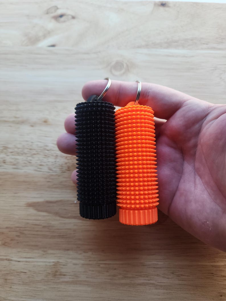 May include: Two textured keychains, one black and one orange, are held up against a wooden background. The keychains have a cylindrical shape with a spiky surface. Each keychain has a silver keyring attached to the top.