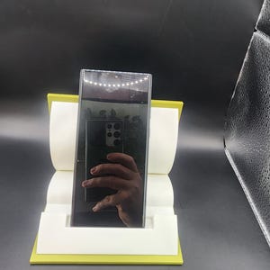 May include: A black smartphone with a camera lens on the back is displayed on a yellow stand. The phone is reflecting the image of a hand.