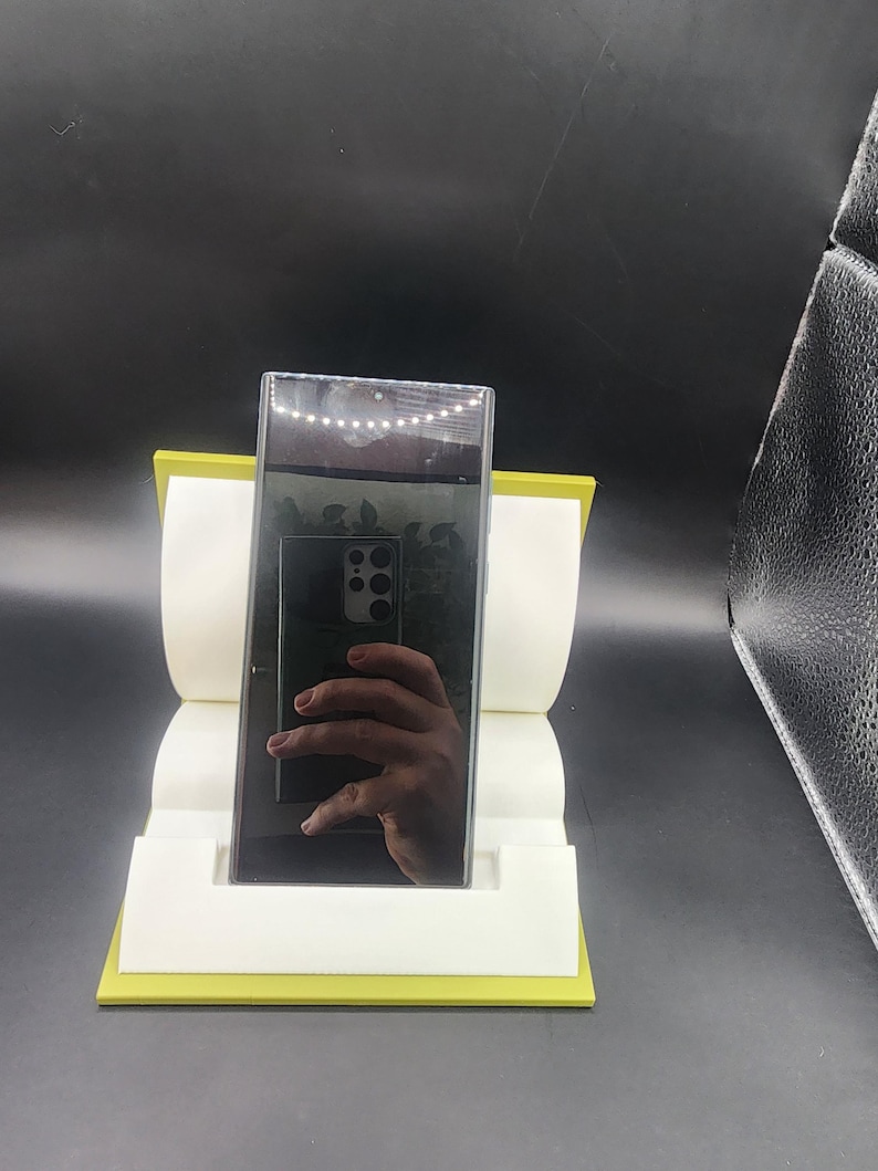 May include: A black smartphone with a camera lens on the back is displayed on a yellow stand. The phone is reflecting the image of a hand.