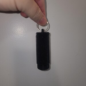 May include: A black, cylindrical keychain accessory with a textured surface. The keychain has a metal ring at the top for attaching keys. The object is held against a plain, light gray background.