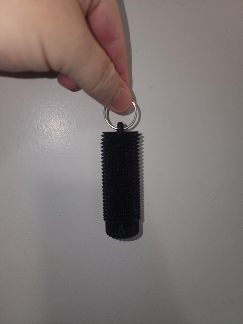 May include: A black, cylindrical keychain accessory with a textured surface. The keychain has a metal ring at the top for attaching keys. The object is held against a plain, light gray background.