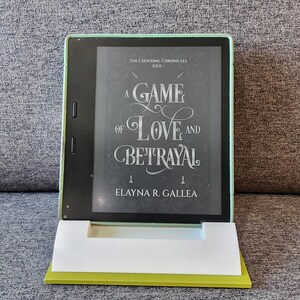May include: A green e-reader displaying the cover of a book titled "A Game of Love and Betrayal" by Elayna R. Gallea. The book is part of a series called "The Choosing Chronicles".