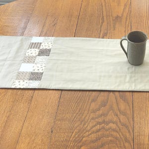 May include: A beige table runner with a patchwork design in shades of brown and white. The runner is on a wooden table with a mug on the right side.