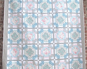 May include: A quilt with a patchwork design featuring a grid of squares in shades of blue, pink, and white. The squares are surrounded by a gray border.