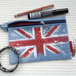 May include: A denim makeup bag featuring a Union Jack design in red and white, with a silver zip and red lining. Also shown are a brown lip pencil, black mascara, a silver bracelet, and feather earrings.