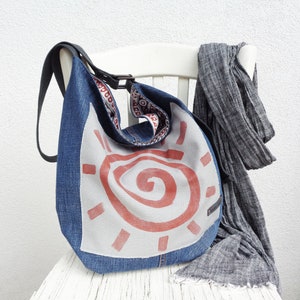 May include: A blue denim shoulder bag with a white panel featuring a red spiral sun design. The bag has a black strap and a patterned interior. A grey scarf hangs on a white chair next to the bag.