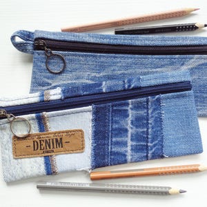 Op de afbeelding: Twee denim etuis met donkerblauwe ritsen en een bruin leren label met de tekst "DENIM". Er liggen verschillende kleurpotloden verspreid rond de etuis. De etuis zijn gemaakt van gerecycled denim.