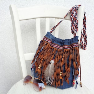 May include: A blue denim bag with brown fringe and a feather accent. The bag has a braided strap and wooden beads. The bag is sitting on a white chair.