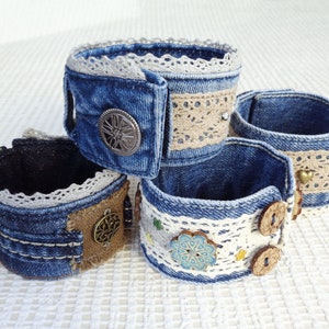 Puede incluir: Cuatro pulseras de puño de mezclilla con adornos únicos. Cada pulsera está hecha de mezclilla azul y presenta ribetes de encaje, botones decorativos y detalles metálicos. Una tiene un diseño floral, otra un emblema metálico.