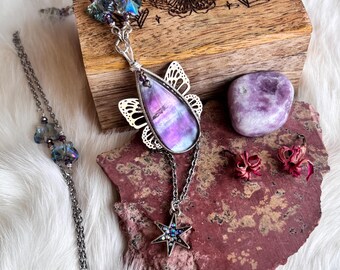 May include: A silver necklace with a large, teardrop-shaped pendant featuring a purple and white stone, accented by a butterfly-shaped filigree. A smaller star pendant hangs below. A wooden box with a butterfly and crystal design is in the background.