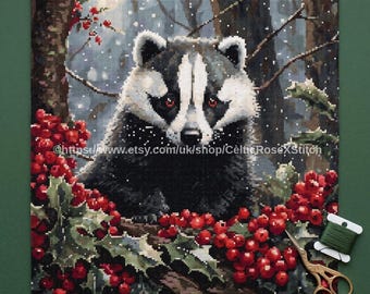 May include: A PDF digital download cross stitch pattern featuring a badger in a snowy forest setting with red holly berries. The image includes a spool of green thread and gold-colored scissors. The text reads PDF Digital Download Cross Stitch Pattern.