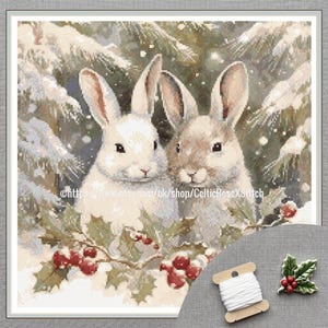 May include: Cross-stitch picture of two rabbits in a snowy scene with holly and red berries. The rabbits have white and brown fur. A spool of white thread and a holly leaf embellishment are in the lower right corner.