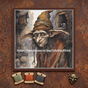 May include: A framed cross-stitch artwork depicts a whimsical goblin character with a long nose, pointed ears, and a mischievous grin. The goblin wears a brown hat and robe, set against a detailed, aged town backdrop. The artwork is signed with a shop URL.
