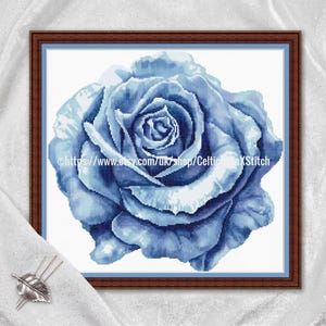 May include: A framed cross-stitch of a blue rose. The rose is in full bloom, with detailed shading creating depth and dimension. The artwork is set against a white background and framed in a dark brown wooden frame. The Etsy shop URL is visible in the image.