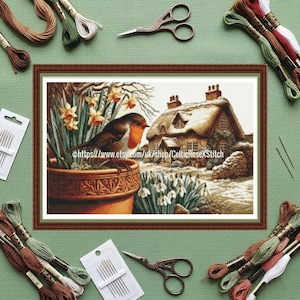 Può includere: Un ricamo a punto croce incorniciato di una scena di cottage con un uccello su un vaso di fiori. La scena comprende un cottage con tetto di paglia, narcisi e bucaneve. Filo da ricamo, aghi e forbici circondano la cornice.