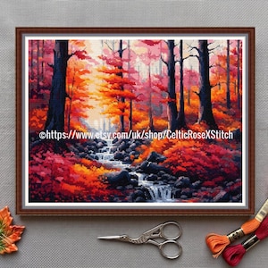 Könnte beinhalten: Gerahmte Kreuzstich-Landschaft eines Herbstwaldes mit einem Bach. Das Kunstwerk verwendet leuchtende Farben von Rot, Orange und Gelb, mit dunklen Baumstämmen und einem fließenden Bach. Ein blattförmiges Ornament und Stickgarn sind ebenfalls vorhanden.