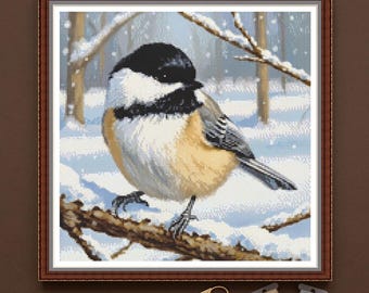 May include: A PDF digital download cross stitch pattern featuring a black-capped chickadee perched on a snow-covered branch. The bird has a black head, white cheeks, and tan underparts. Below the framed pattern are gold scissors and two spools of thread.