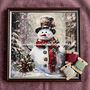 May include: Framed cross-stitch art of a snowman in a winter forest scene. The snowman wears a top hat, red scarf, and has a carrot nose. The frame is brown. Spools of thread are in the foreground.