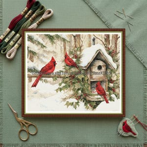 Peut inclure: Une broderie au point de croix encadrée représentant une scène hivernale avec des cardinaux rouges perchés près d'un nichoir enneigé. L'œuvre est dans un cadre marron, avec du fil à broder et des ciseaux visibles. Le texte "www.etsy.com/uk/shop/LilacRoseXStitch" est inclus.
