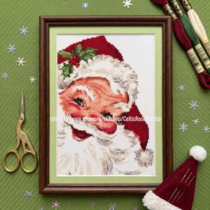May include: Framed cross-stitch of Santa Claus with a red hat and white beard. The image is surrounded by embroidery floss, a small Santa hat, gold scissors, and snowflake decorations. The Etsy shop URL is visible on the frame.