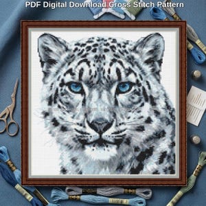 May include: A PDF digital download cross stitch pattern featuring a detailed portrait of a snow leopard with striking blue eyes, black spots, and white fur, presented within a brown frame against a textured blue fabric background.