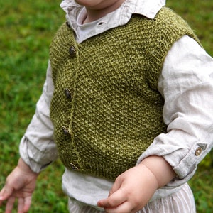 May include: A young child wearing a green knitted vest with metal buttons over a white shirt with a light brown striped pants.