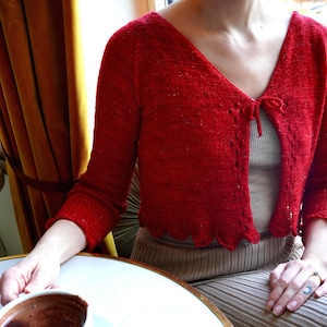 May include: A woman wearing a red knitted cardigan with a tie front and a beige skirt, sitting at a table with a cup of hot chocolate.