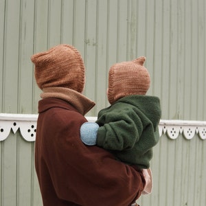 May include: A person wearing a brown hooded hat holds a child wearing a green hooded jacket and a brown hat. The child is wearing brown shoes.