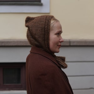 Op de afbeelding: Een vrouw draagt ​​een bruine gebreide muts en een bruine jas. De muts heeft een lange flap die de nek bedekt.