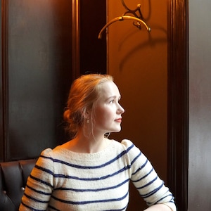 May include: A woman wearing a white and blue striped sweater sits at a table with a cup of coffee. She is looking out the window.