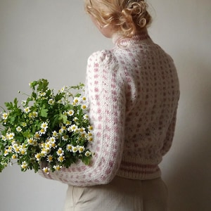 May include: A woman wearing a pink and white patterned jumper holds a bouquet of white daisies and green foliage.