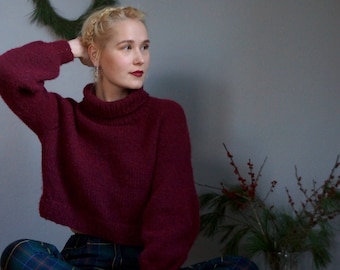 May include: A woman wearing a burgundy turtleneck sweater and plaid pants sits with her legs crossed. She has blonde hair styled in a braid and is looking to the side.