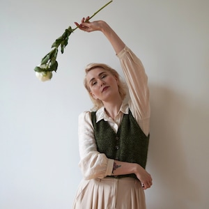 May include: A woman wearing a white shirt, a green knitted vest, and a beige skirt holds a single white flower with green leaves.