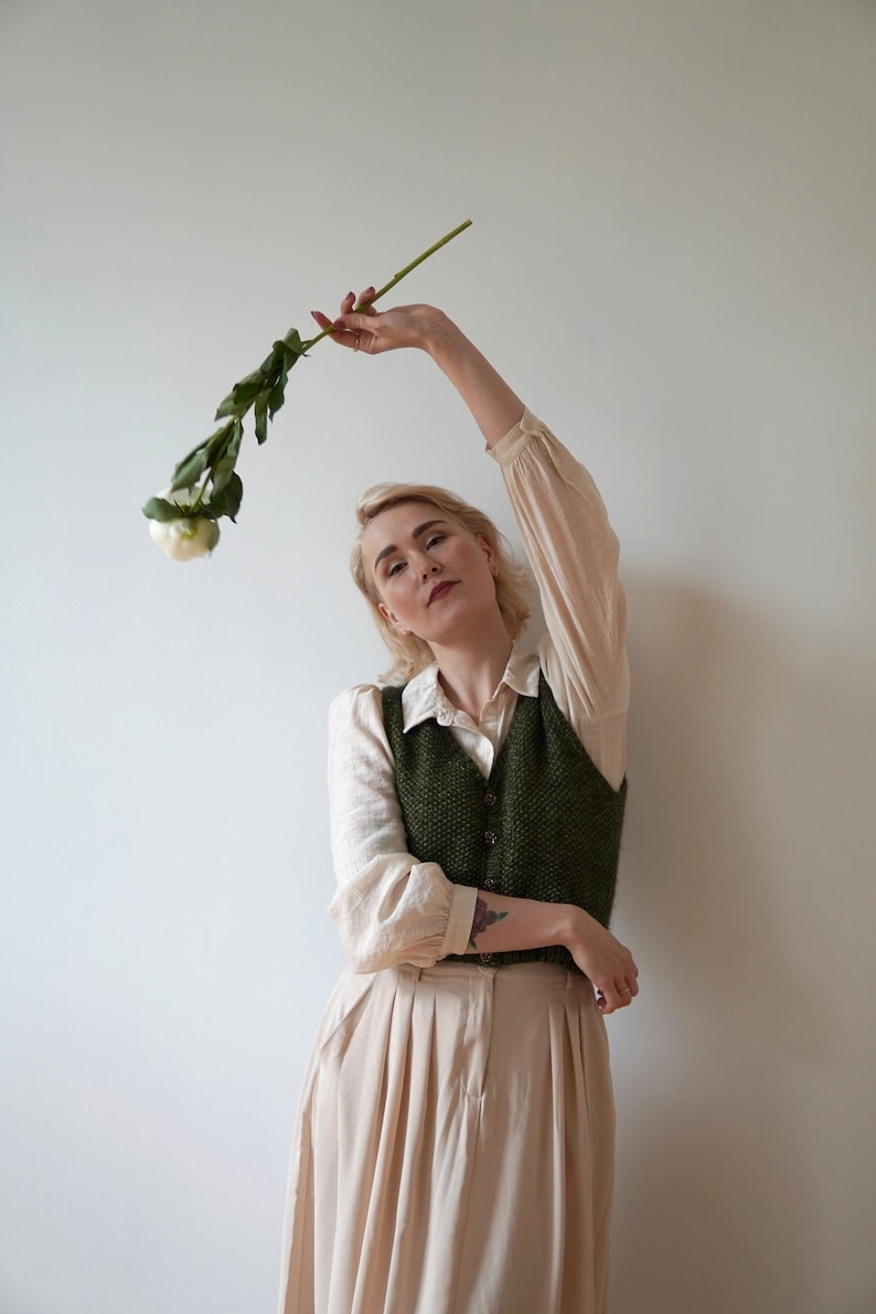 May include: A woman wearing a white shirt, a green knitted vest, and a beige skirt holds a single white flower with green leaves.