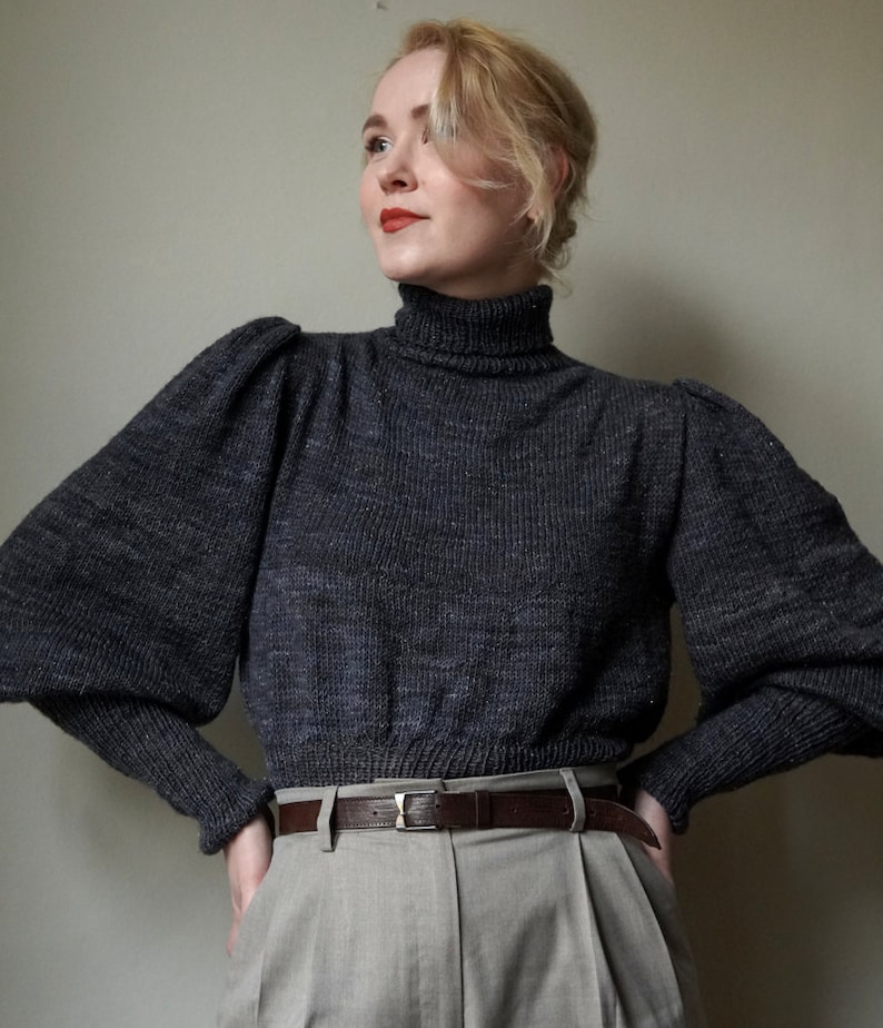 May include: A woman wearing a dark gray turtleneck sweater with puffed sleeves and a brown leather belt. She is wearing beige pants.
