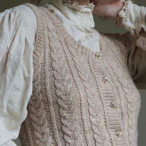 Op de afbeelding: Een beige kabelgebreid vest met V-hals, geribbelde zoom en vijf decoratieve knopen met een bloemachtig middendetail. Het kledingstuk wordt gedragen over een crèmekleurig shirt met lange mouwen, een ruchekraag en manchetten met knopen.