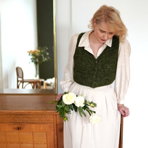 May include: A woman wearing a white shirt and a green knitted vest holds a bouquet of white peonies. She is standing in front of a wooden cabinet with a mirror in the background.