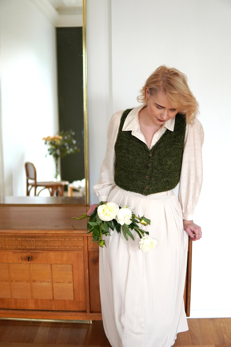 May include: A woman wearing a white shirt and a green knitted vest holds a bouquet of white peonies. She is standing in front of a wooden cabinet with a mirror in the background.