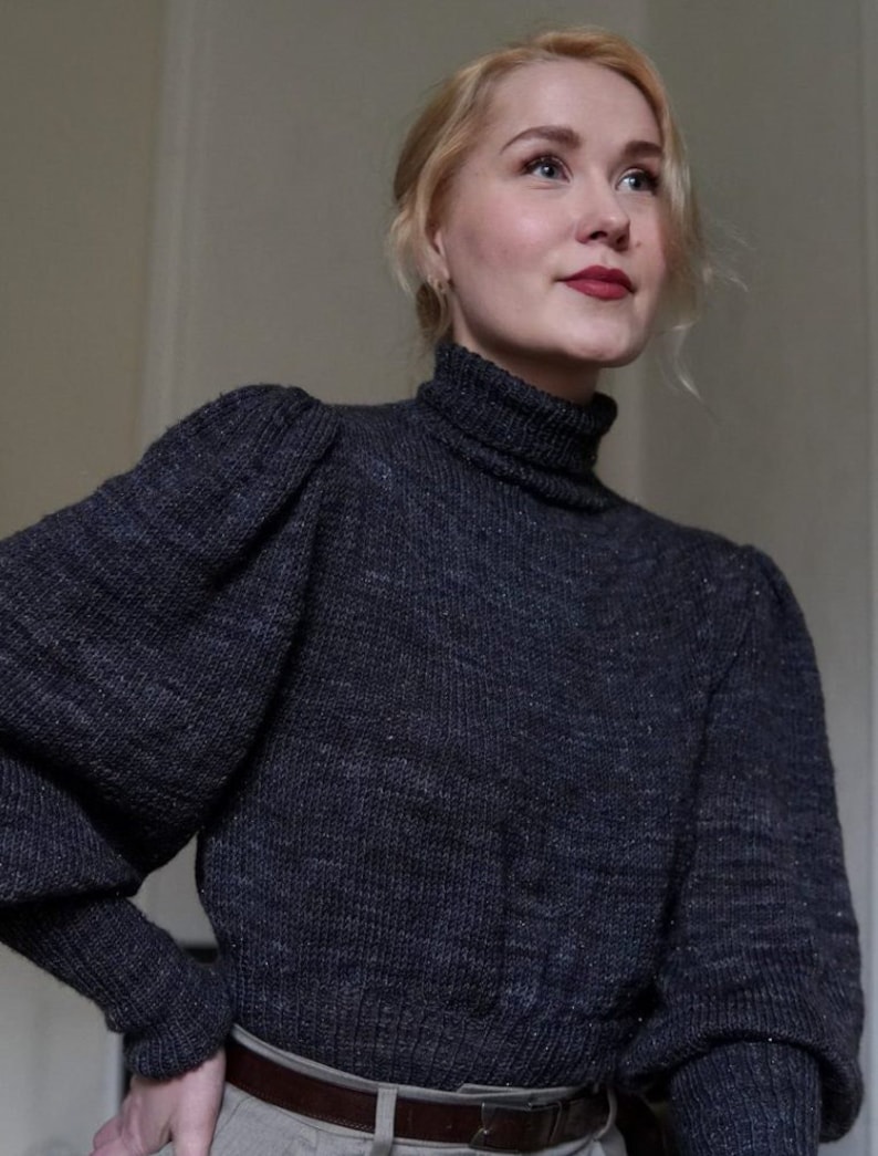 May include: A woman wearing a dark gray turtleneck sweater with puffed sleeves. The sweater has a subtle sparkle and is worn with a brown belt.