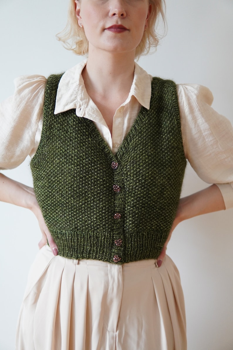 May include: A green knitted vest with a V-neck and button closure. The vest is made of a textured yarn and has a casual, relaxed fit.