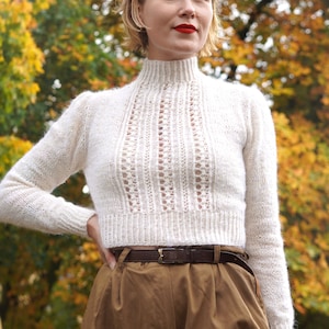 Puede incluir: Una mujer con un suéter blanco de punto con un delicado patrón calado y pantalones marrones de cintura alta. Está de pie frente a un fondo borroso de hojas de otoño.