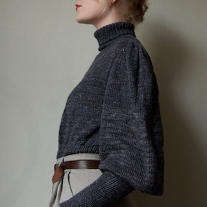 May include: A woman wearing a dark gray turtleneck sweater with puffed sleeves and a brown belt. She is wearing light gray pants.