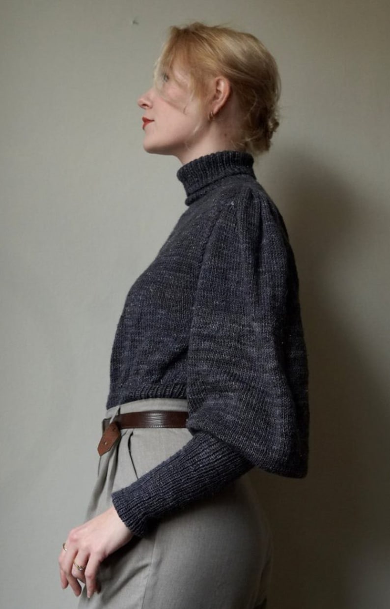 May include: A woman wearing a dark gray turtleneck sweater with puffed sleeves and a brown belt. She is wearing light gray pants.