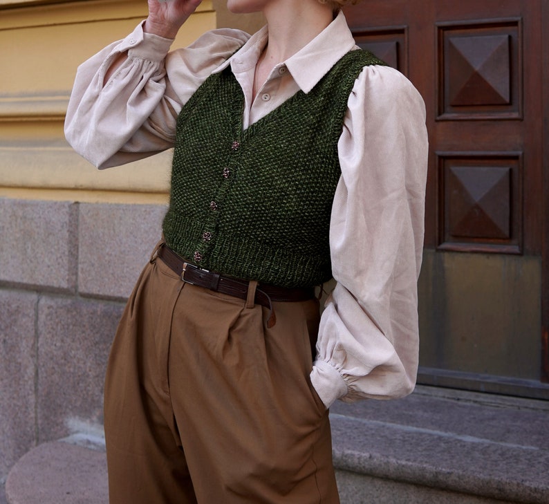 May include: A green knitted vest with a white button-down shirt underneath. The vest has a V-neck and is worn over brown pants with a brown leather belt.