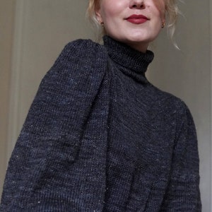 May include: A woman wearing a dark gray turtleneck sweater with puffed sleeves. The sweater has a subtle sparkle throughout the fabric.