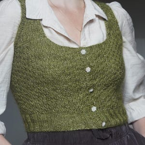 May include: A sleeveless olive green knit vest featuring a square neckline and a front button closure with five small, round, silver-toned floral-shaped buttons. The vest has a textured stitch pattern and a ribbed hem at the waistline.