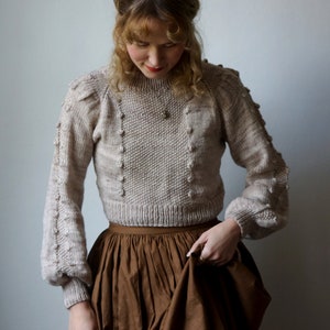 Puede incluir: Una mujer lleva un suéter de punto beige con una falda marrón. El suéter tiene un patrón texturizado y un escote redondo. La falda es completa y está fruncida en la cintura.