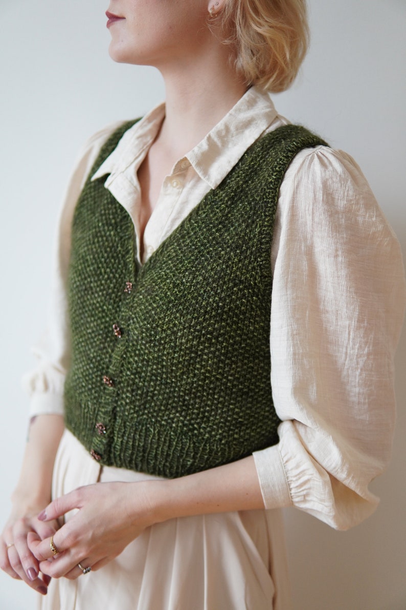May include: A green knitted vest with a V-neck and button closure. The vest is worn over a white button-down shirt.