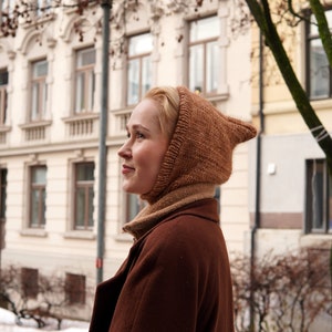 Könnte beinhalten: Eine Frau in einer braunen gestrickten Mütze mit Kapuze und einem braunen Mantel, die vor einem Gebäude mit Schnee auf dem Boden steht.