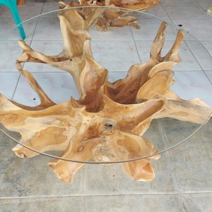 May include: A unique coffee table featuring a clear, round glass top supported by a natural, light brown, intricately carved wooden base. The base has a root-like appearance with multiple branches and a textured surface. The table's design is organic and modern.